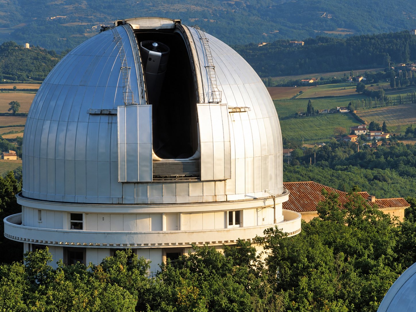 Grande coupole métallique de l’observatoire de Saint-Michel, fente ouverte, entourée d’arbres avec des champs et des collines en arrière-plan.