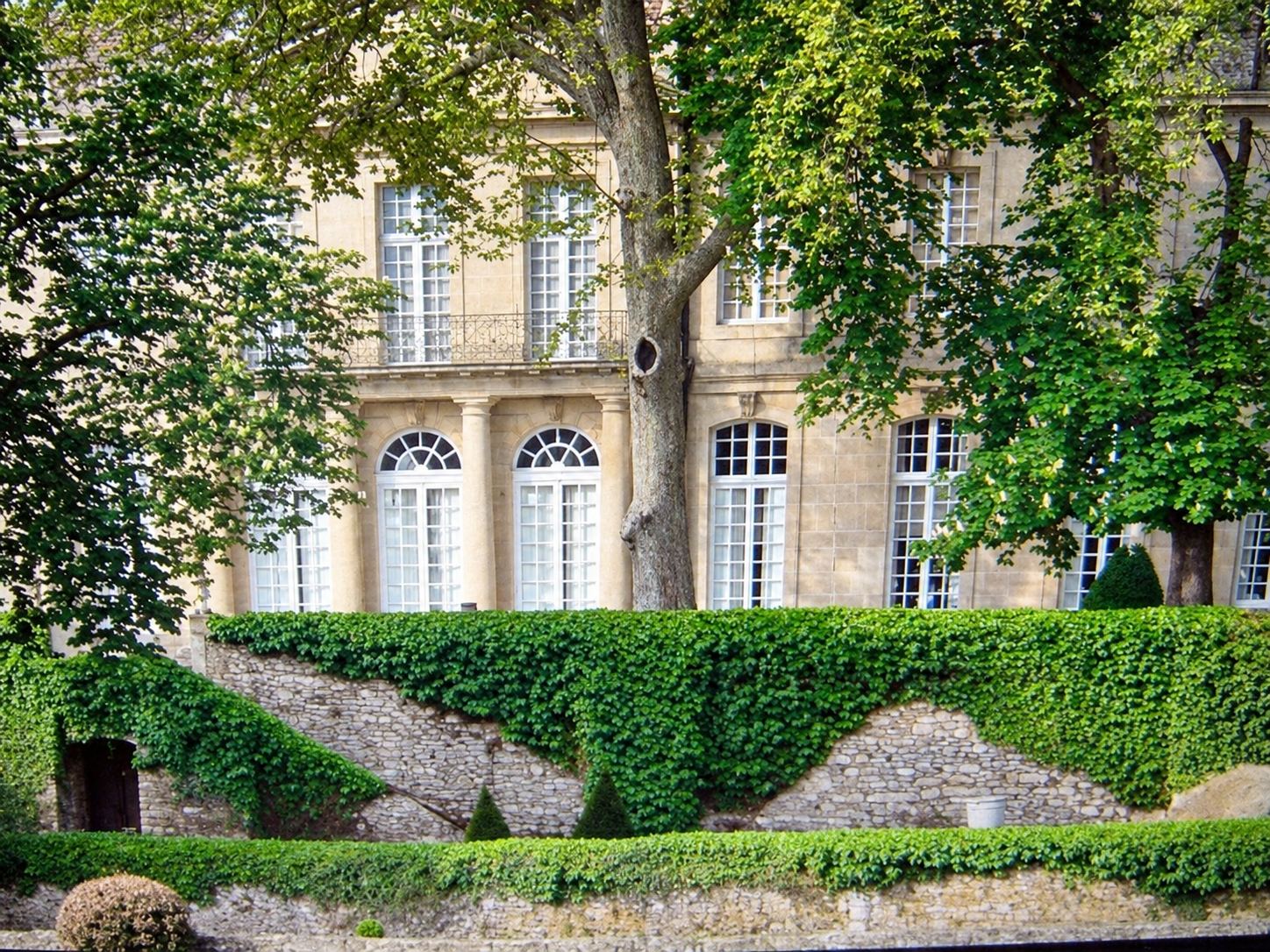 Façade en pierre claire du château de Sauvan, aux grandes fenêtres, entourée d’arbres et d’un mur de pierre couvert de lierre.