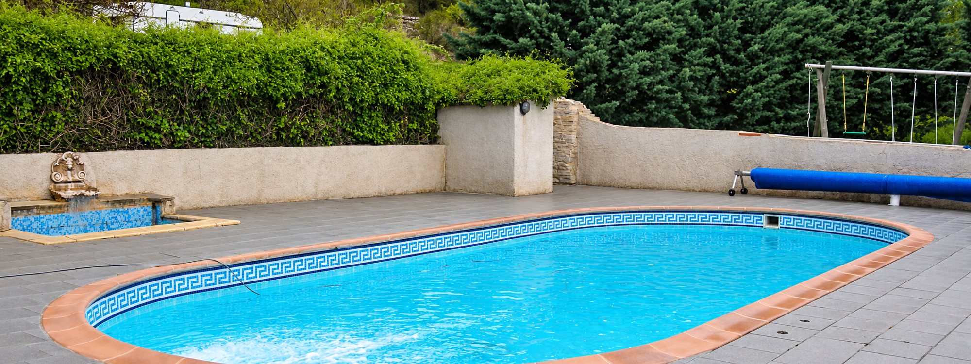 Piscine ovale à l’eau turquoise bordée de margelles orangées et d’une frise bleue à motif grec, avec terrasse dallée, murs en pierre et haies.